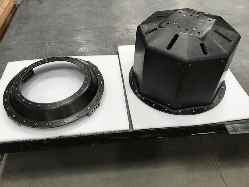 Image shows the two pieces of the smallsat launch adapter structure in a manufacturing area and looks down at them from above and to the side. They are sitting on a dark-colored pallette with a protective white foam layer on its surface. The structural ring is to the left and the octagonal structure is on the right.